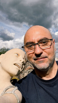Close-up portrait of a man posing together with an artistic mannequin sculpture outdoors on a sunny day with scattered clouds.