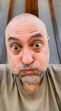 A close-up selfie of a middle-aged bald man with a beard making a funny face with wide eyes and pursed lips against a textured glass background.