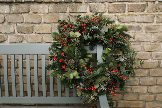 Wooden bench decorated with Beautiful Handmade Christmas wreath. Evergreen Foliage, Red Berries, Ivy, Eucalyptus. Elegant advent porch decoration. Blurred brick background. Winter outdoor home decor