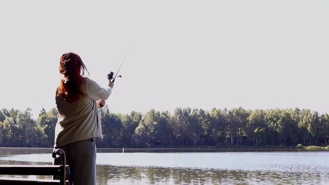 Wide shot of a red-haired female angler fishing with a spinning rod on a wooden pier near a river or lake on a sunny autumn day. Concept of outdoor activities, hobby, and relaxation in nature.