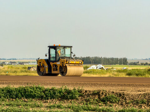 Saskachewan, Canada - August 20, 2026: Caterpillar CB16 tandem vibratory roller compacting road base on flat prairie construction site with farmland and wind turbines in background