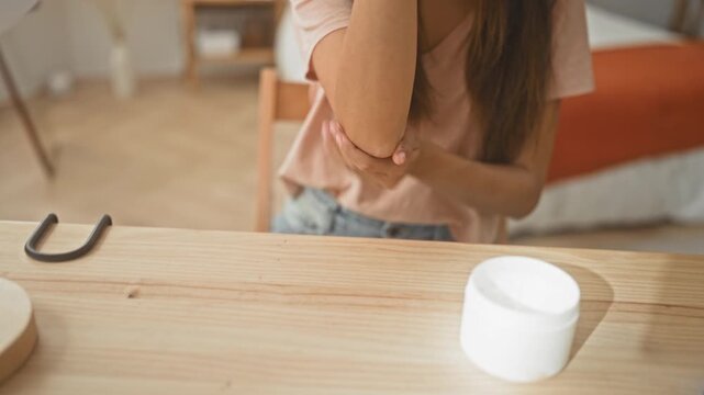 Young hispanic woman rubbing lotion on elbow and clapping hands over open cream jar on a wooden table in a studio; self care relaxation.