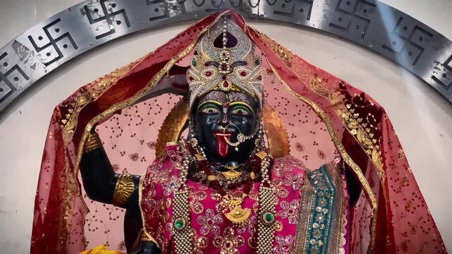 A close-up devotional view of Maa Kali featuring a distinctive dark face, ornate crown, layered jewelry, and vibrant red and pink ceremonial drapery.