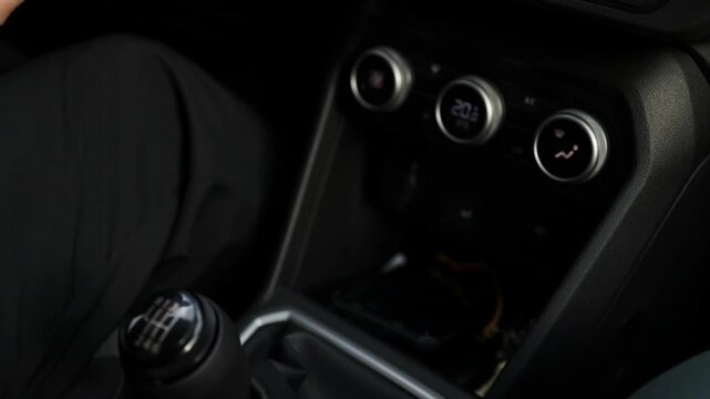 Close up video frame of a driver's hand holding a manual transmission gear stick lever on the center console. In the background, the modern car dashboard displays illuminated round climate control