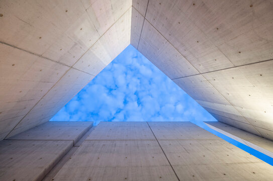 Interior view of a board-formed concrete structure with a triangular opening revealing a blue sky with clouds. 