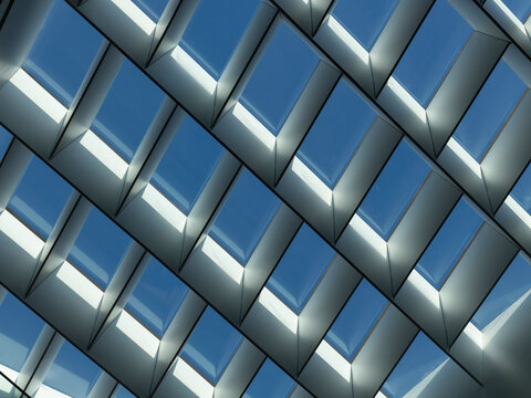 Close-up view of a skylight roof structure formed by a repeating diamond grid of angled metal fins against a blue sky. 