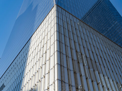 Low-angle view of a business office tower in Lower Manhattan, where a corner of textured metal fin cladding meets a smooth glass curtain wall, both rising against a clear blue sky.