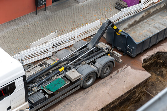 Hooklift truck loads broken asphalt and concrete into roll off container during urban roadwork. Excavator, rubble and heavy machinery show demolition, recycling and infrastructure repair process