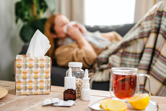 Sick woman resting on a sofa with tissues, while tea, lemon, capsules, nasal sprays and medicine sit on a table. Cold symptoms, seasonal illness, recovery and self care.