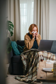 Woman working on a laptop while recovering from a cold at home, wrapped in a blanket and using a tissue, with tea and medicine nearby. Illness and remote work.