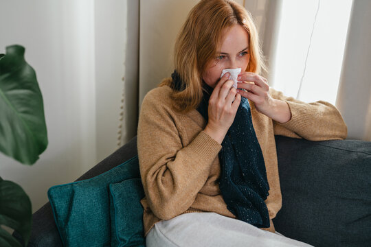 Woman blowing her nose with a tissue while sitting on a sofa at home. Cold, flu, seasonal allergies, illness, recovery and self care