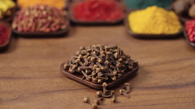 dried cloves buds in a square bowl on a wooden table, surrounded by various spices and herbs.