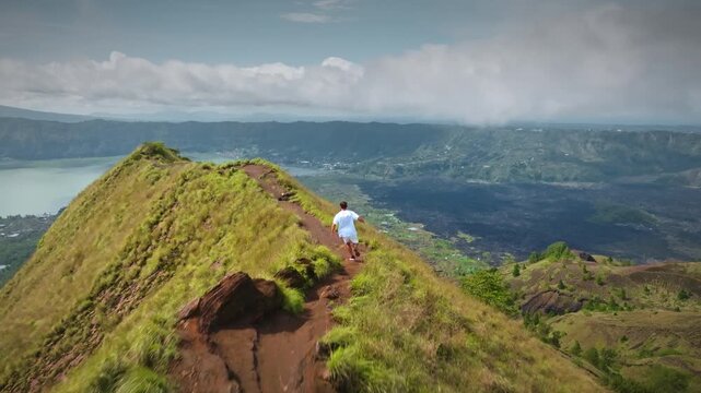 Person hiking running on grassy ridge of Mount Batur dark volcanic caldera, Lake Batur, and lush green landscape under cloudy sky, active sports, adventure and exploration. Aerial tracking drone shot