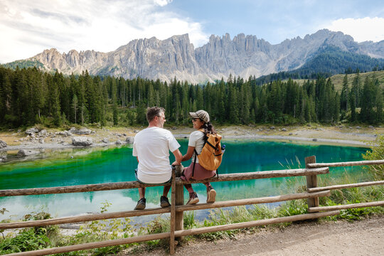 A couple sits on a wooden fence, gazing into each others eyes, surrounded by the breathtaking landscapes of Lago di Carezza in the Dolomites, Italy.