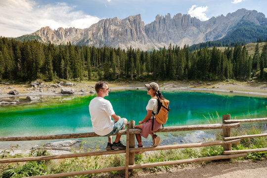 A couple relaxes on a wooden fence by the stunning Lago di Carezza in the Dolomites, Italy. The clear turquoise water reflects the majestic mountains and lush pine trees, inviting serenity and peace.