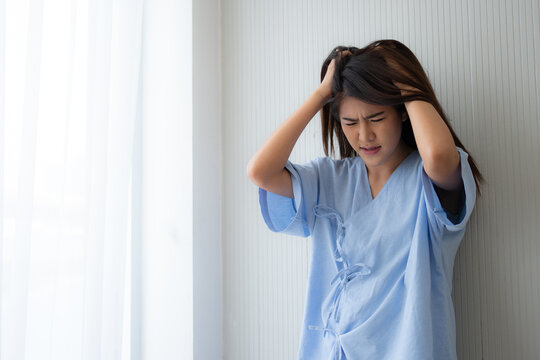 Young woman hospital patient with an IV line holds her head in pain while sitting in bed, conveying severe headache, illness, emotional distress, recovery, healthcare treatment and clinical care.