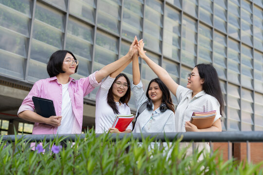 Diverse female college students celebrate outdoors beside a modern university building, raising their hands and holding books to represent education, teamwork, friendship, unity, and achievement.