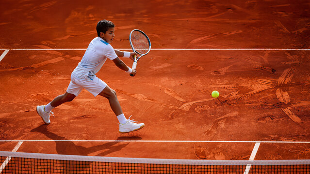 Young Indian boy playing tennis hitting backhand shot on clay court during training match action