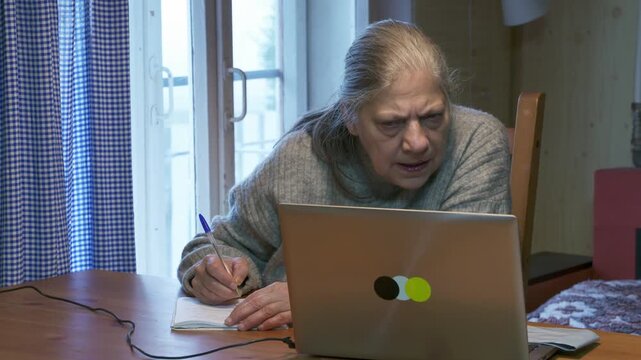 An older woman writes notes in a notebook while looking at a laptop computer at a wooden table indoors. Her focused and diligent study session feels calm and peaceful.
