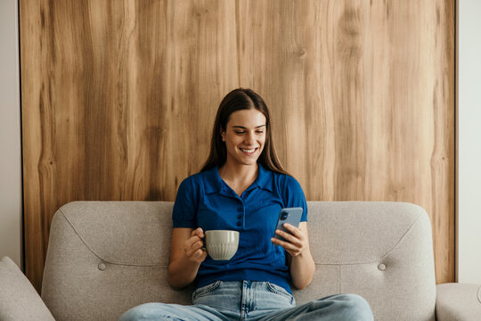 Woman relaxing on sofa using smartphone and drinking coffee