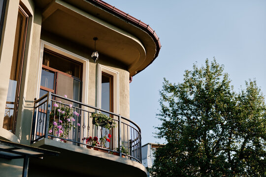 Cozy house balcony with curved architecture, metal railing and blooming flowers, green tree and blue sky in background. Concept of home comfort and residential lifestyle.