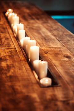 Glowing white pillar candles arranged inside a long central recess of a handcrafted wooden table, creating warm ambient light across richly grained timber with soft selective focus