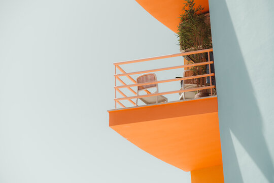 Minimal architectural detail of a bright orange balcony with cane chairs and a potted plant projecting from a pale blue wall, framed by clean geometric lines and sunlight