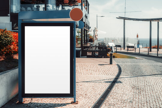 Blank vertical advertising board in a dark freestanding frame outside a waterfront restaurant, ready for branding or menu design with an ocean promenade visible beyond