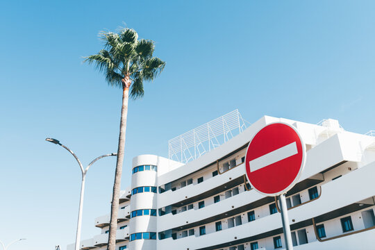 Red no entry traffic sign stands against a tall palm tree and curved white hotel architecture beneath an expansive clear blue sky in bright summer sunlight