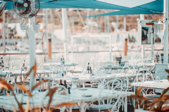 Empty coastal dining terrace with ornate white chairs and marble tables beneath blue shade sails beside a yacht marina, prepared for guests on a bright summer day; shallow depth of field