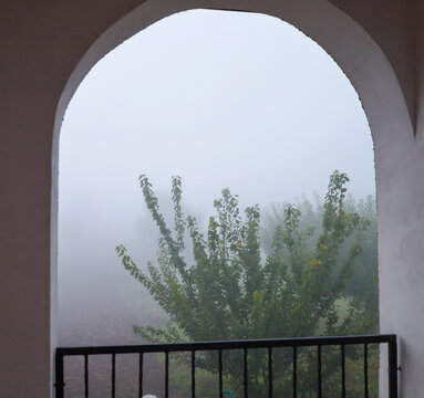 View from a balcony or terrace through a white arched opening into thick autumn fog. Green crowns of trees and bushes peep through the misty haze, creating an atmosphere of mystery and tranquility.