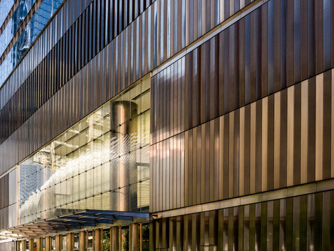 Exterior view of a business office building in Manhattan clad in vertical bronze-toned metal fins, with a glass canopy entrance below reflecting a curved column and interior lighting. 