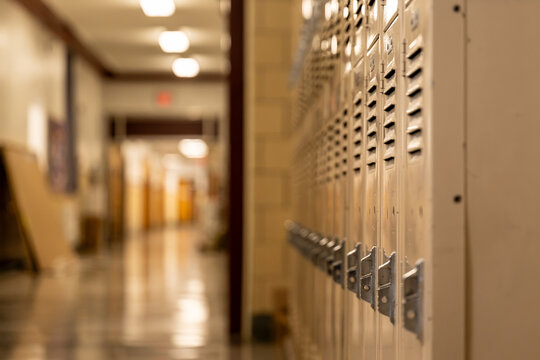 An empty school hallway recedes past lockers and wooden doors beneath ceiling lights, evoking back to school, student absence, transition, public education, and campus life.