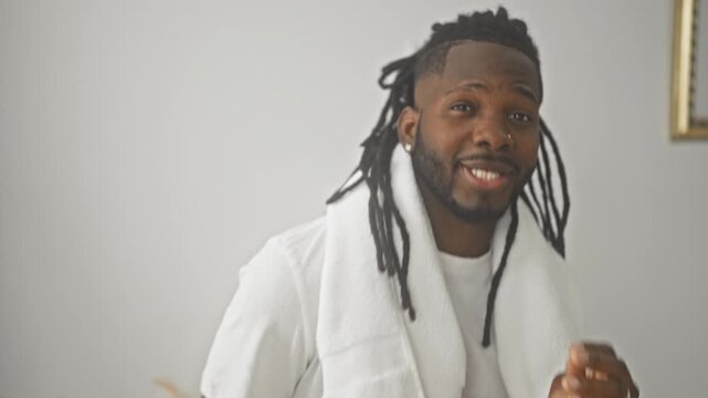 Young african american man with dreadlocks draped in towel touching face with hand, wearing white tshirt in studio; grooming relaxed.