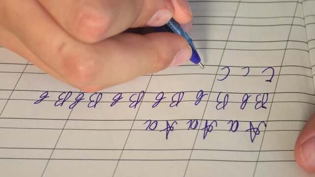 Close-up of a hand holding a blue pen writing cursive uppercase and lowercase letter С on grid-lined notebook paper