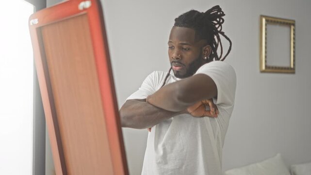 Young african american man crosses arms at full length mirror in studio, wearing white tshirt, dreadlocks, nose stud and chain, gold frame on wall; contemplation.