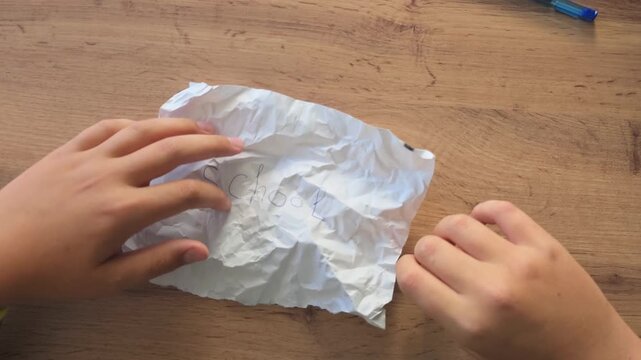 Two hands holding a crumpled white paper sheet with faint cursive writing; wooden table surface in background, blue pen visible