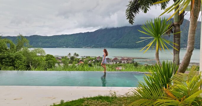 Young beautiful woman walking on the edge of an infinity pool in a modern villa resort, overlooking Lake Batur and Mount Batur in Bali, enjoying stunning volcanic landscape and serene natural scenery