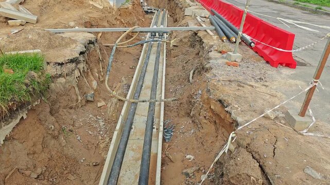 Construction trench with underground cable conduits in concrete trays; exposed earth walls, red plastic barrier, scattered construction debris, utility pipes and clamps
