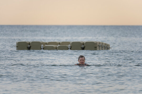 Langrune-Sur-Mer, France - 08 09 2026: A mature woman wearing a blue swimsuit is swimming in the sea with a swimming platform behind