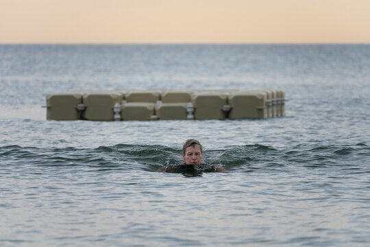 Langrune-Sur-Mer, France - 08 09 2026: A mature woman wearing a blue swimsuit is swimming in the sea with a swimming platform behind