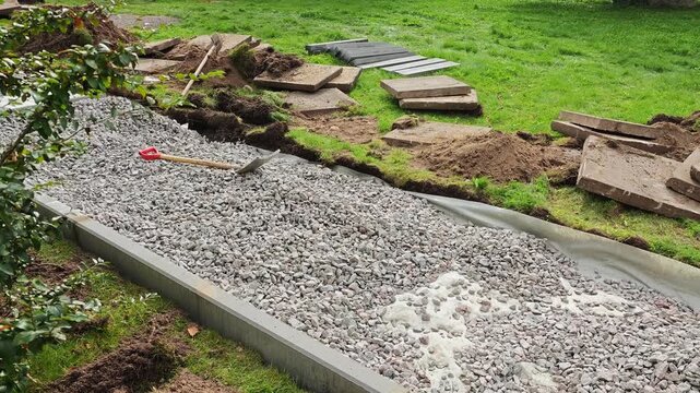 Construction site for a gravel pathway: concrete border, geotextile layer, loose gravel fill, scattered soil piles, concrete slabs, nearby shrub, shovel on gravel, green lawn background.