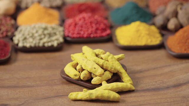 dried turmeric root or curcuma longa in a square bowl on a wooden table, surrounded by various spices and herbs.