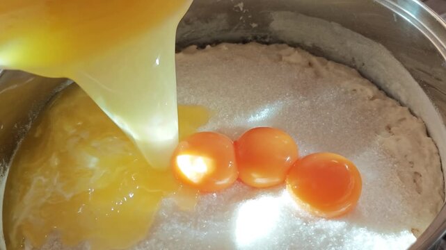 Golden egg yolks and egg whites pouring from a bowl into a stainless steel pot with white granular batter. Close-up cooking process, baking ingredients.