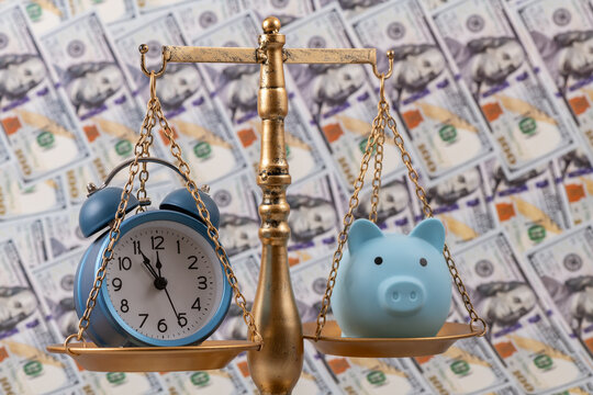 A piggy bank and an alarm clock on the scales against a background of US $100 bills.