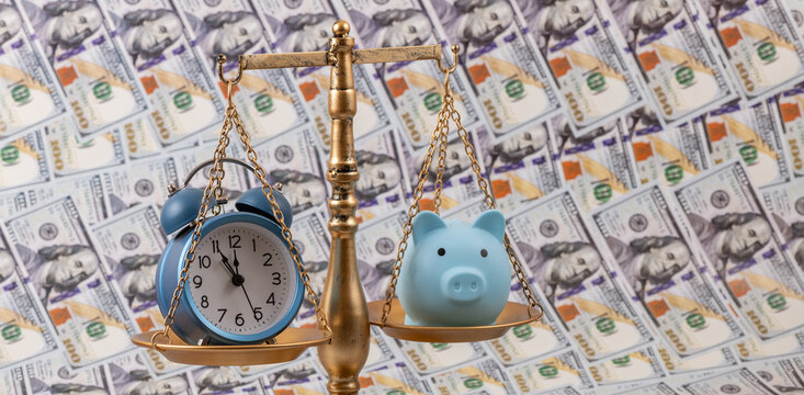 A piggy bank and an alarm clock on the scales against a background of US $100 bills.