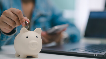 Naklejka na ściany i meble Young male freelancer putting coin into piggy bank working with laptop computer and mobile phone for personal finance savings and investment concept.
