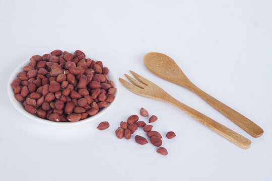 Raw peanuts in a white bowl with wooden spoon and fork isolated on white background. Healthy food ingredient and organic snack concept.