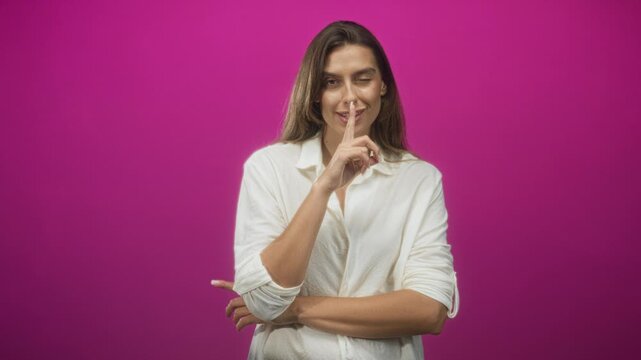 Young hispanic woman finger to lips for silence and folded arms in studio with magenta backdrop; playful secrecy.