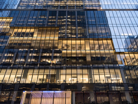 Wide exterior view of a business office building in Manhattan at dusk, its glass curtain wall revealing multiple illuminated interior floors of open workspaces.